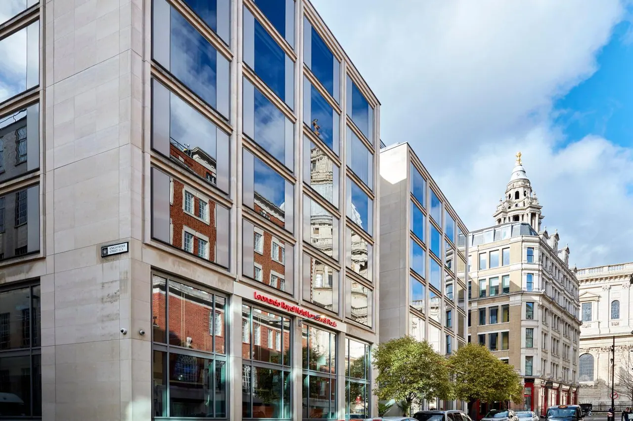 The exterior of a pale stone and glass hotel building with red Leonardo Royal Hotel London St Paul's signage at street level, with an ornate historic tower and part of St Paul's Cathedral visible to the right.