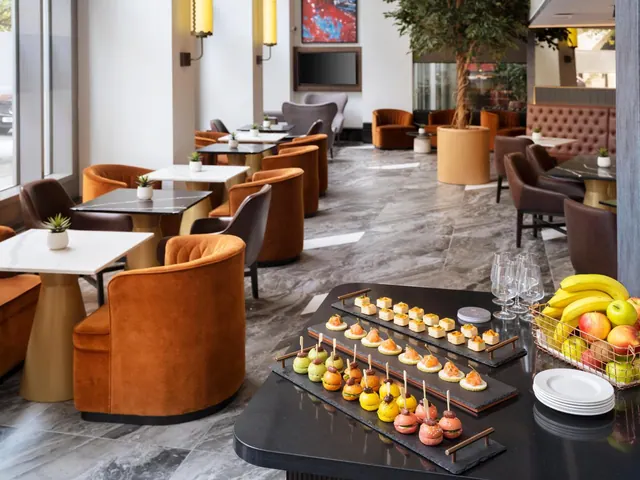 A lounge area with orange velvet armchairs and marble tables, with a buffet of canapés and a fruit basket on a dark table in the foreground.