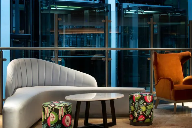 A lounge seating area with a curved grey sofa, two patterned stools, a round marble table and an orange armchair, with glass lifts behind a glass balustrade.