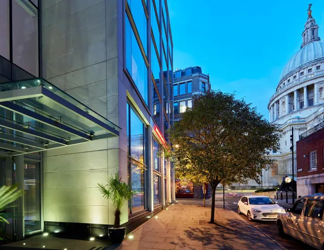 Exterior of a glass and stone hotel building with a covered entrance canopy, a street tree and parked cars, with the dome of St Paul's Cathedral visible on the right at dusk.