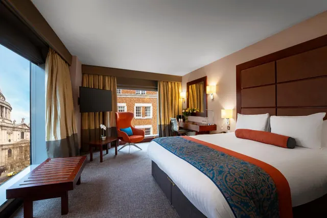 A hotel bedroom with a king bed, an orange armchair, a desk, a wall-mounted television and a large window showing St Paul's Cathedral.