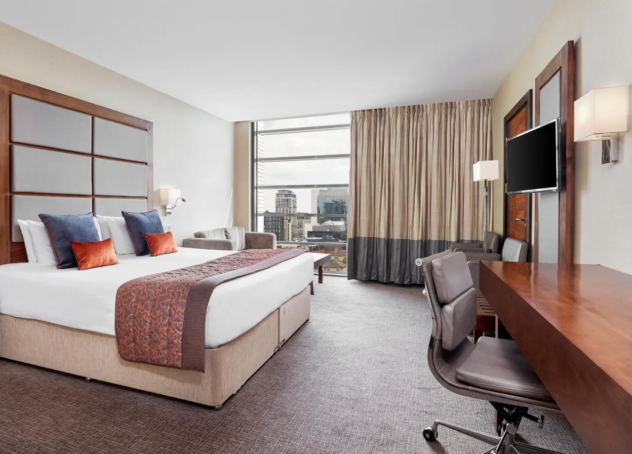 A hotel bedroom with a king bed, a small sofa, a wooden work desk with a wall-mounted television, and a floor-to-ceiling window showing city buildings.
