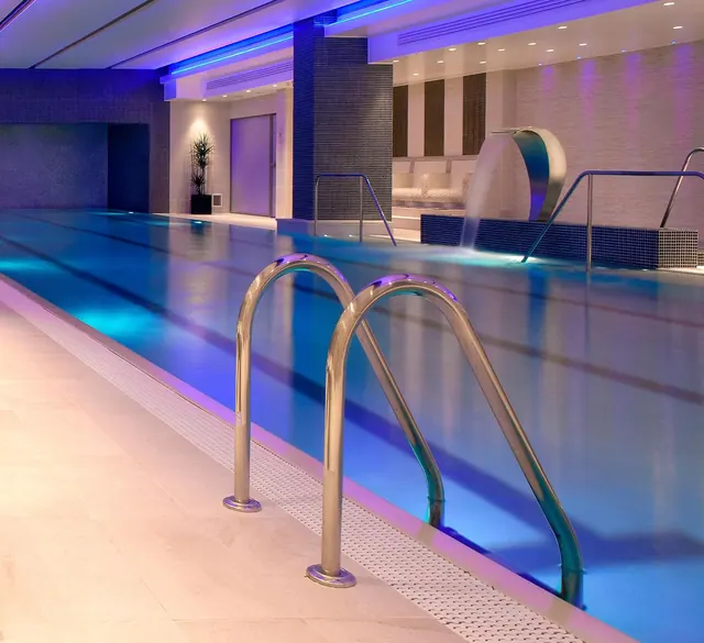 An indoor swimming pool with stainless steel handrails in the foreground, blue lighting, and a curved water feature spilling into the pool at the back.