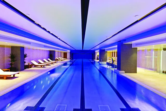An indoor lap pool with a blue-lit ceiling and tiled pillars, with sun loungers lined along one side and a smaller pool area on the right.