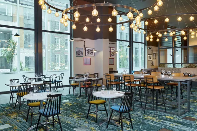 A restaurant dining room with pendant bulb lights, wooden and black chairs at round and high tables, and large windows looking onto a brick building.