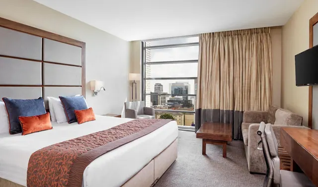 A hotel bedroom with a large bed, a sofa and armchairs, a desk and a wall-mounted television, with a window looking out over city buildings.