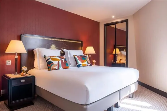A bright hotel bedroom has a large double bed, patterned cushions, bedside lamps and mirrored wardrobe doors.