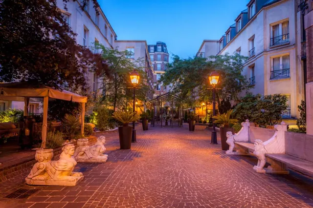 A landscaped hotel courtyard has brick paving, illuminated lamps, ornamental plants, and sculptural benches between surrounding buildings.
