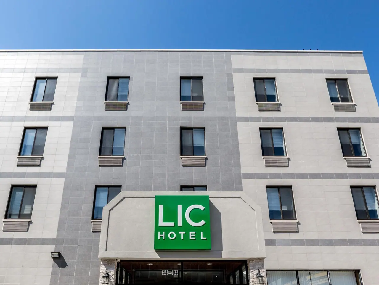 Front facade of the LIC Hotel building, with a green illuminated sign above the entrance and a clear blue sky behind it.