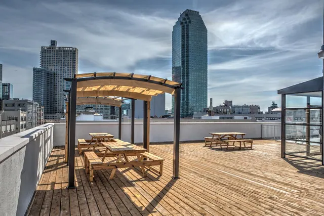 A wooden rooftop terrace with picnic tables under curved canopies, with high-rise buildings visible beyond the parapet.