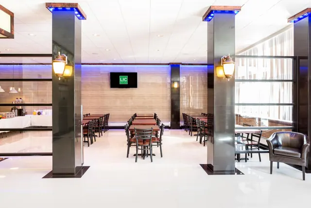 A dining room with wooden tables and chairs, illuminated columns, wall sconces, a wall-mounted TV and a leather armchair beside a large window.