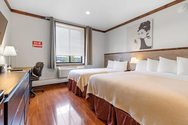 A hotel bedroom with two double beds, a wooden desk and chair, a wall-mounted television, and an air conditioner under the window.