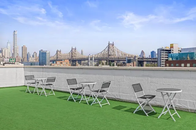 A rooftop terrace with artificial grass and white folding chairs and tables, overlooking the Queensboro Bridge and a Manhattan skyline.