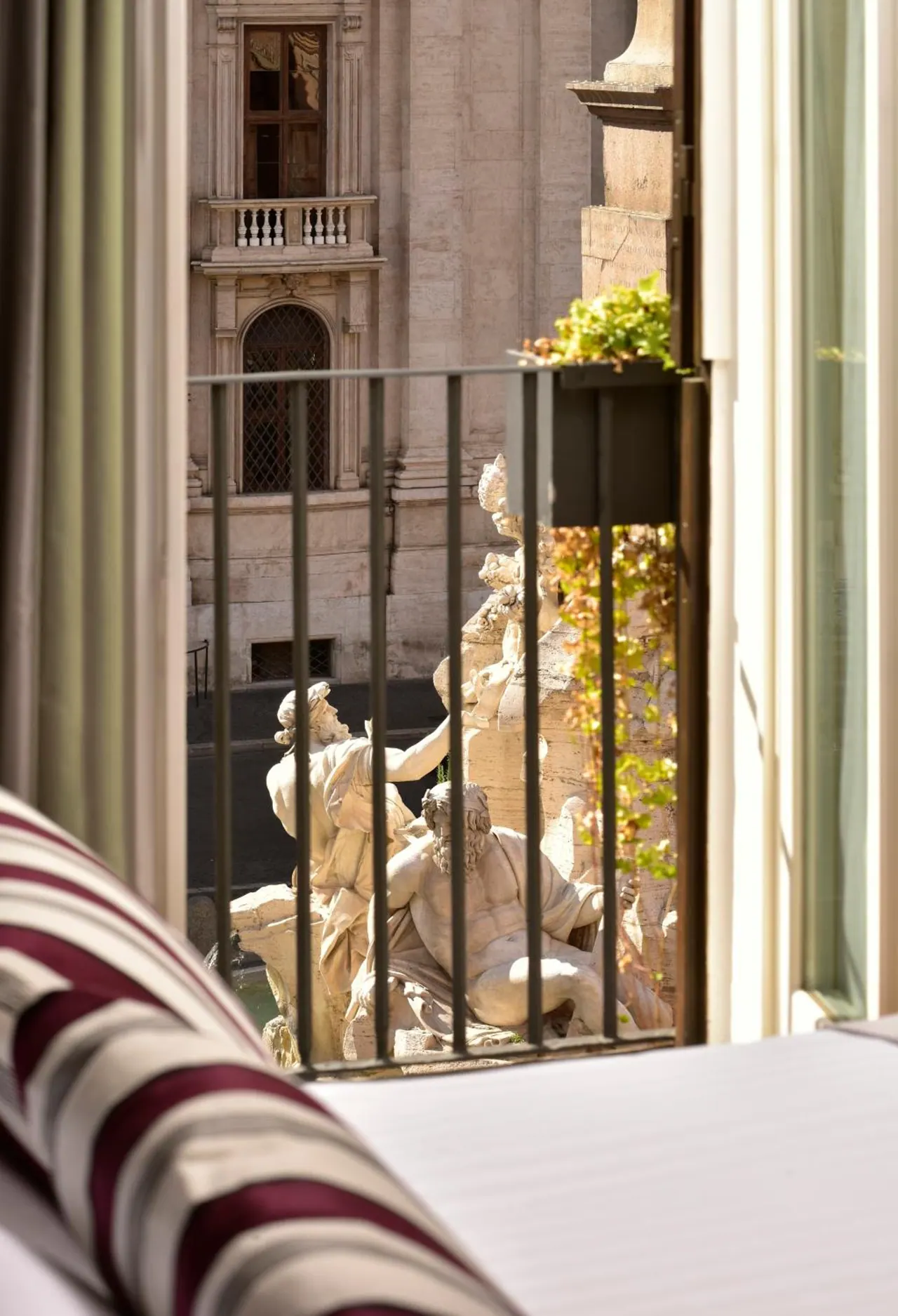 View from a hotel room window through iron railings onto a fountain with marble river-god statues and a historic building facade.