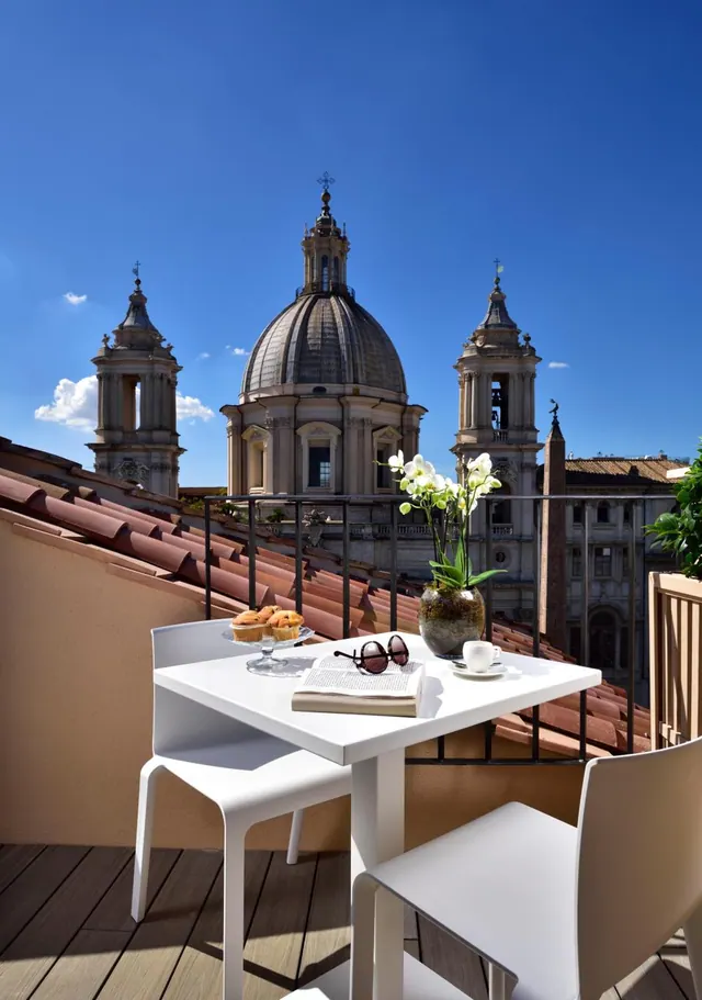 A white table and chairs on a rooftop terrace overlook terracotta roof tiles, with a domed church and bell towers in view under a clear blue sky.