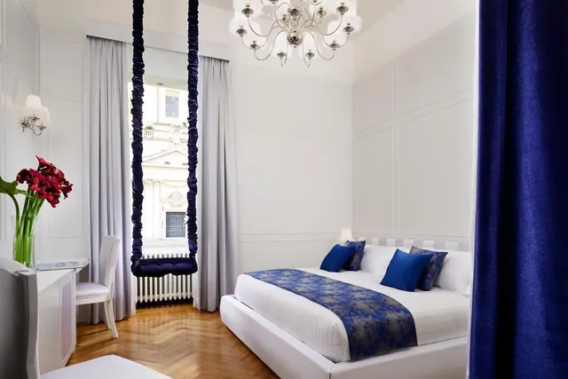 A white bedroom with a made double bed with blue accents, a hanging swing in front of a window, a chandelier, and a white desk with a vase of red flowers.