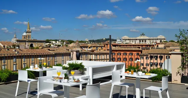 A rooftop terrace with white tables and chairs set for breakfast, overlooking Rome's rooftops, church towers and the white Vittoriano monument under a blue sky.