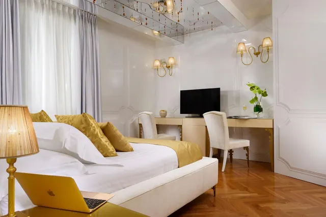 A bedroom with a made white bed with gold accent pillows, a gold laptop and lamp on the bedside table, a wall-mounted television above a desk with two chairs, and gold wall sconces.