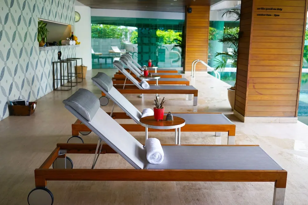 Rows of wooden sun loungers with grey cushions and folded white towels line a covered poolside terrace, with a pool and garden visible beyond the glass at right.