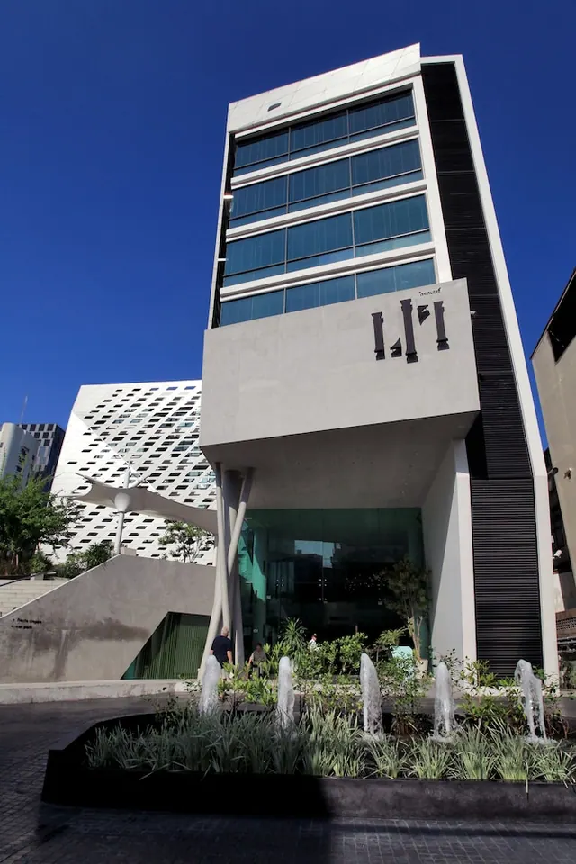 A low-angle view of a tall glass and concrete tower with a large sign reading LI, with fountains and planting in front under a clear blue sky.