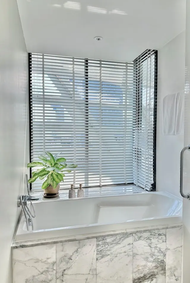 A bathroom with a marble-clad bath beneath a window with closed slatted blinds, a potted plant and two toiletry bottles on the sill.