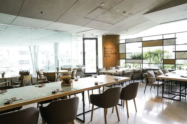 A modern dining room with wooden tables and upholstered chairs, with large windows on the right showing green plants outside.