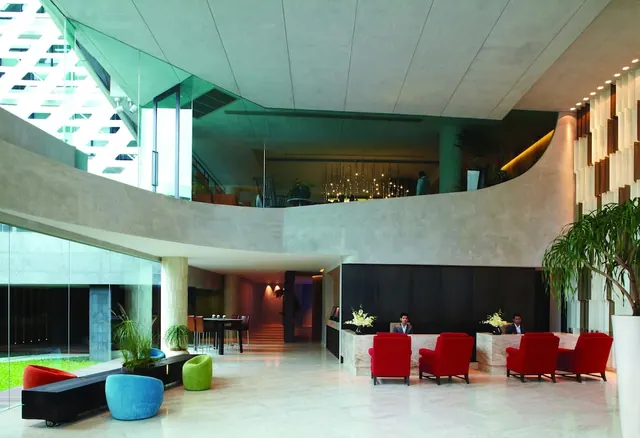 The lobby has red armchairs, coloured pouf seats, a curved upper balcony, and two staff seated behind a reception desk.
