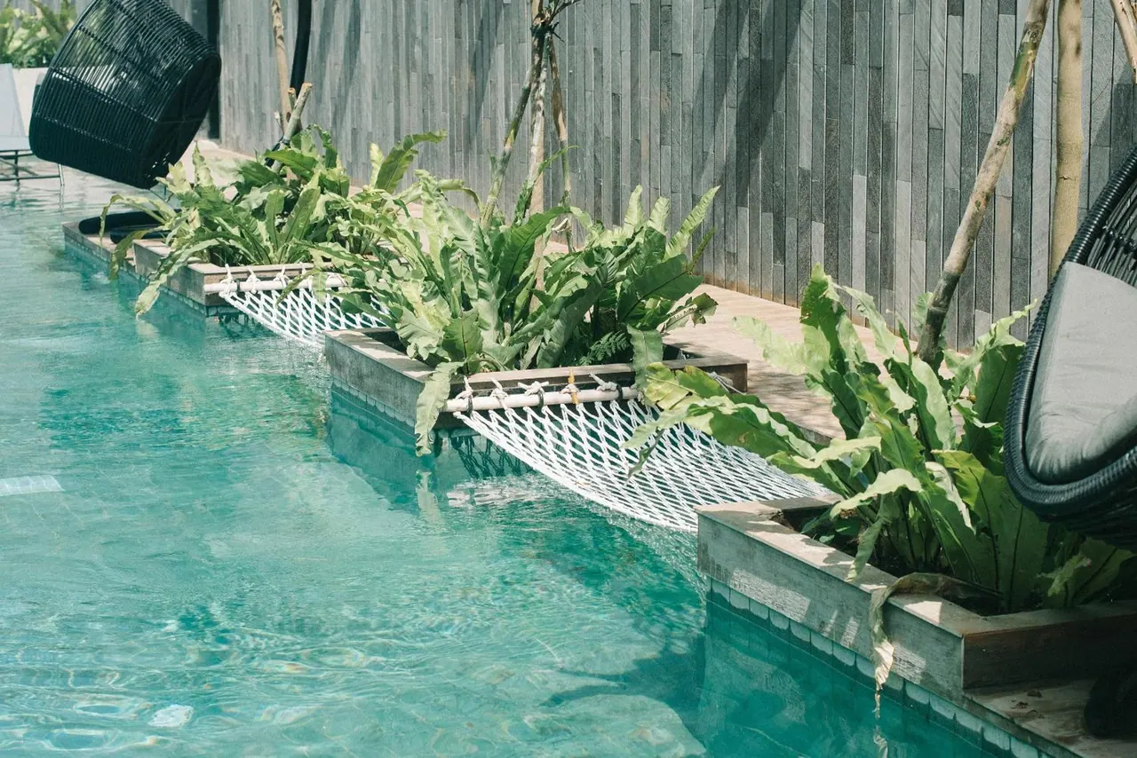 A turquoise swimming pool with fern-filled planters along its edge, black rattan hanging chairs at the corners, and a dark slatted wall behind.