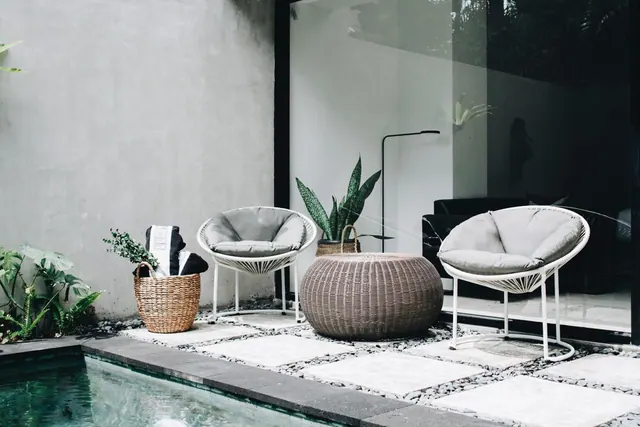 Two white hoop chairs with grey cushions, a wicker side table and potted plants sit on a stone terrace beside a small pool.