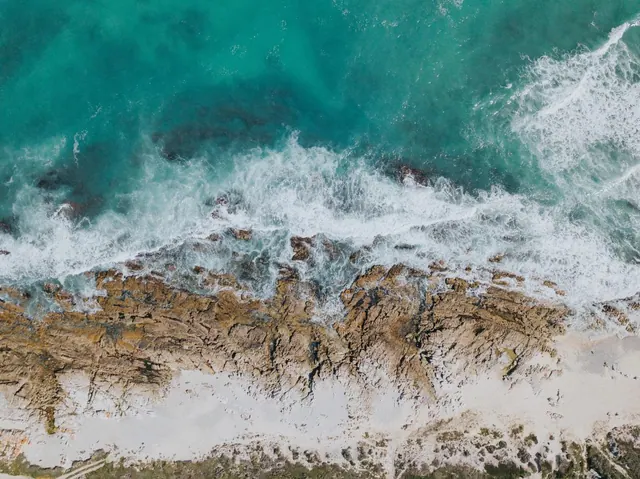 Aerial view of turquoise ocean waves breaking over a rocky shoreline and sandy beach.