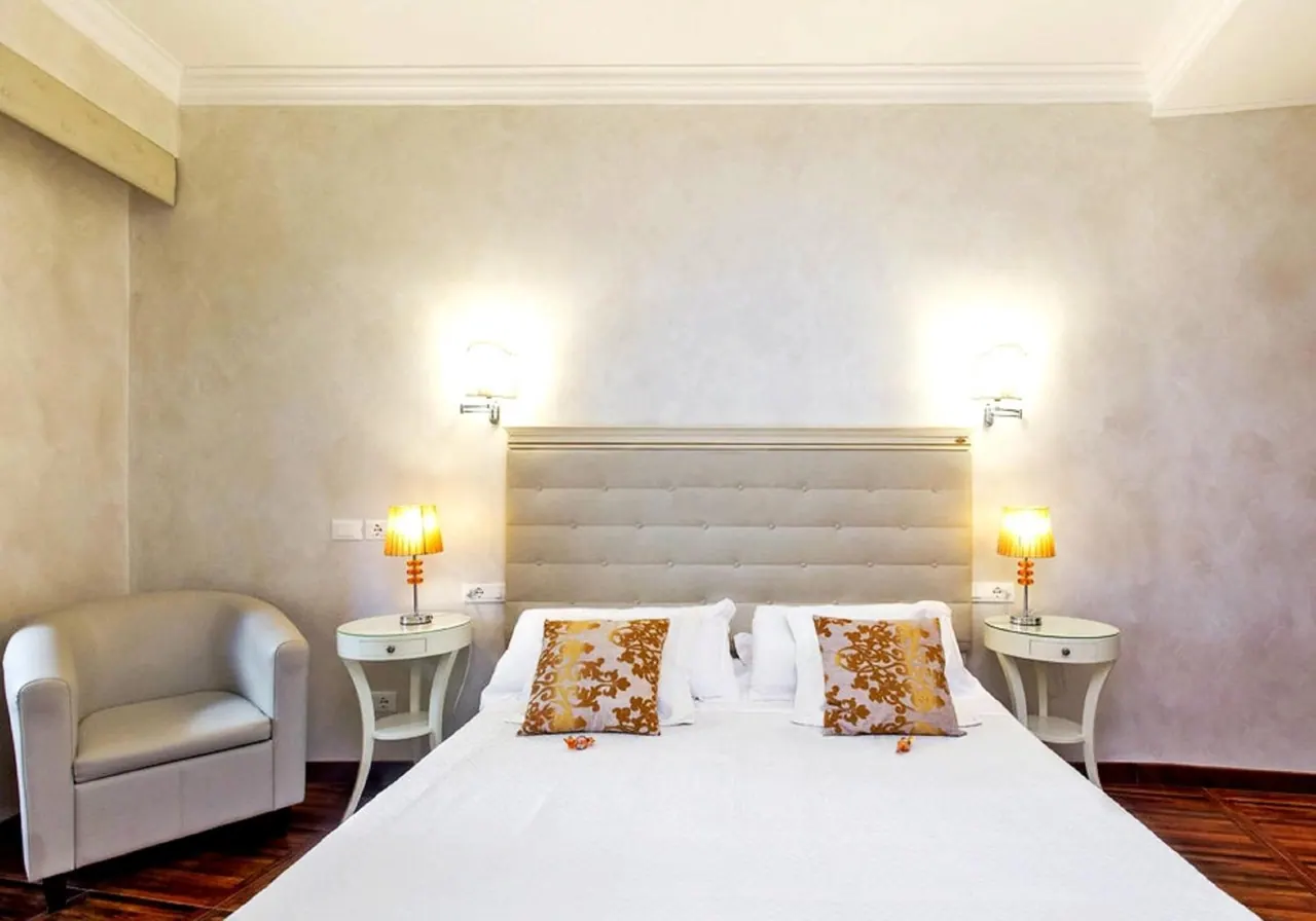 A bedroom with a large white bed, a tufted headboard, two bedside tables with lamps, wall lights, and a white armchair on a wooden floor.