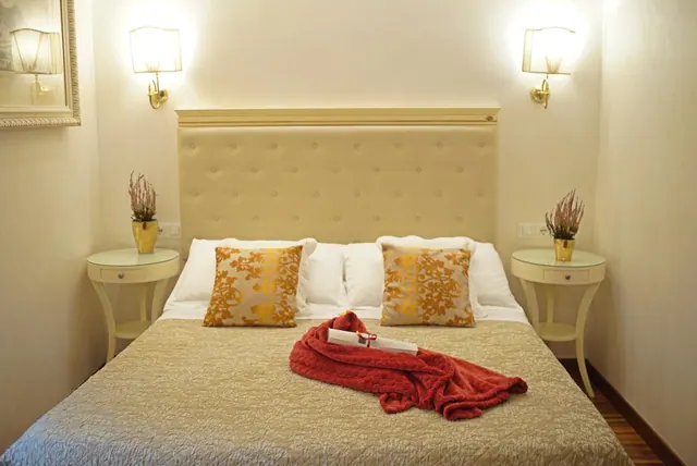 A double bed with a tufted cream headboard, gold patterned cushions and a red throw with a small box on the cover, flanked by bedside tables with potted plants and wall lamps.
