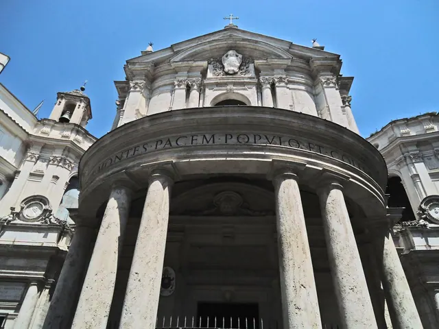 Looking up at the curved stone facade of a baroque church, with tall columns, an inscription band, a carved relief and a cross against a clear blue sky.