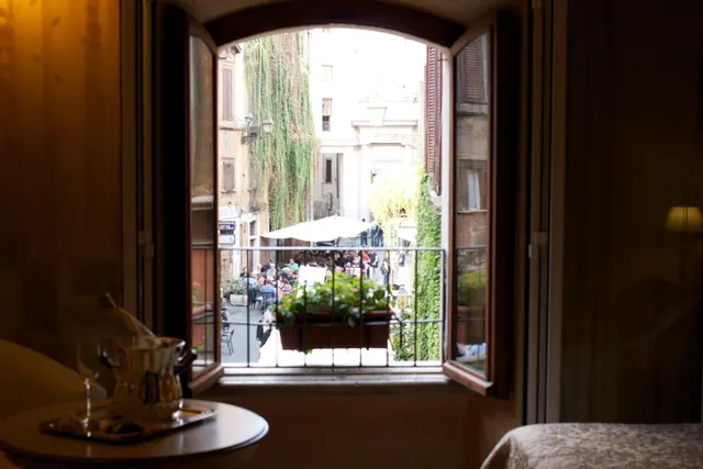An arched window with open shutters frames a view of a street with outdoor café tables and people, a planted window box, and a foreground table holding an ice bucket and glasses.