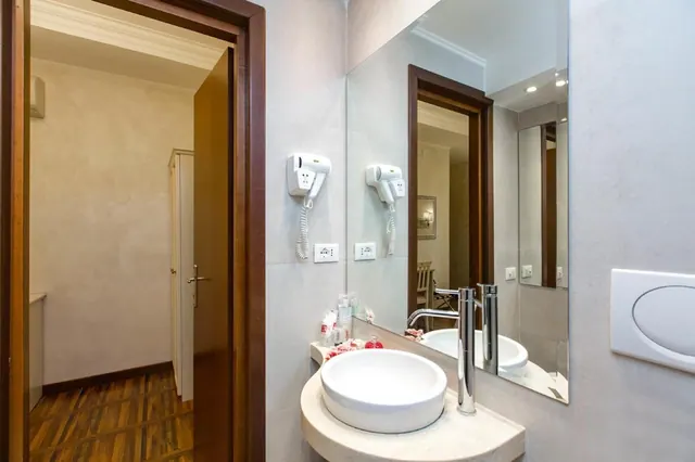 A bathroom with a round white basin on a stone counter, a wall-mounted hair dryer, a large mirror, and an open door leading to a room with a wooden parquet floor.