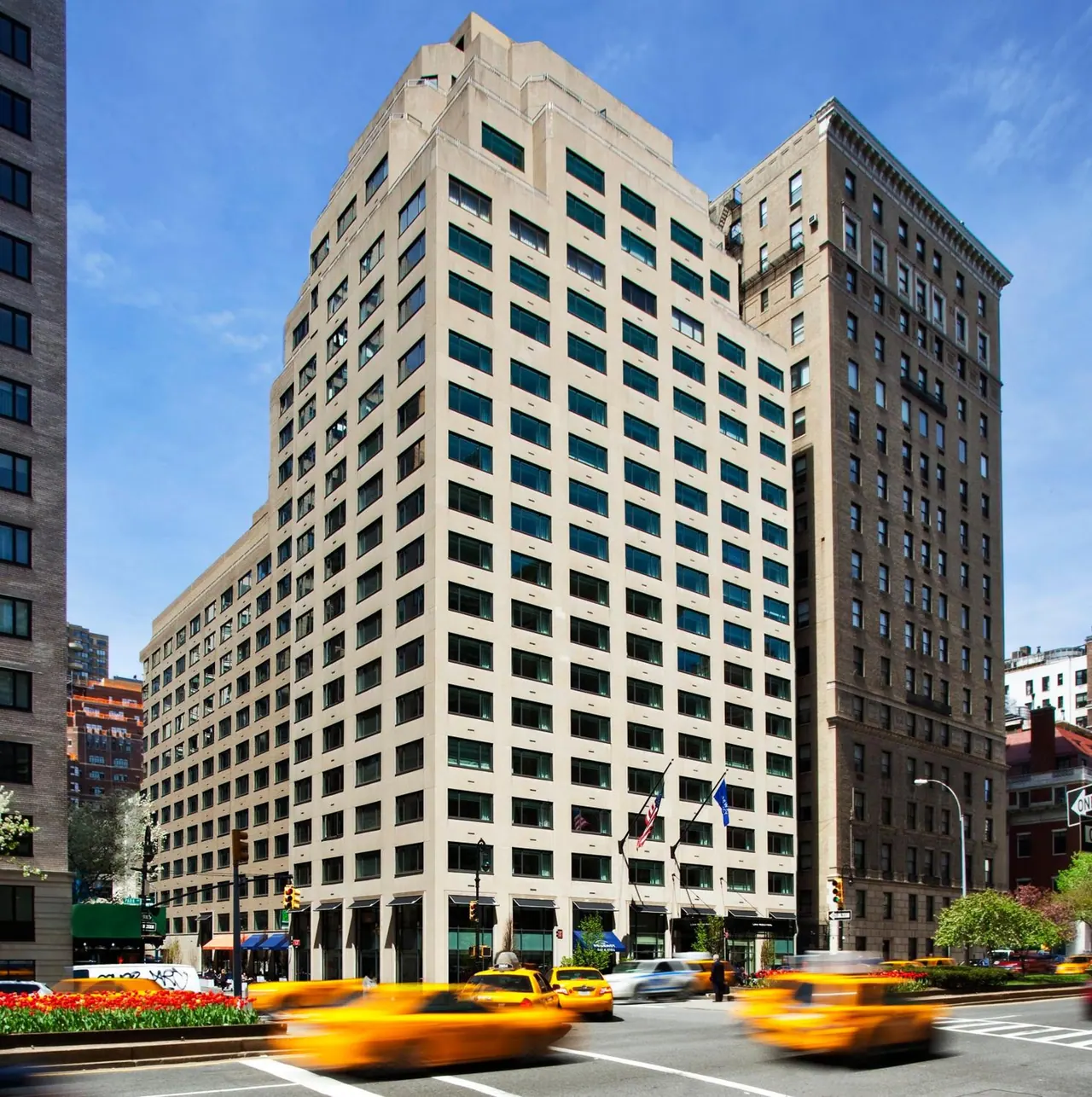 The photo shows the exterior of a tall beige building with rows of windows, with blurred yellow taxis passing on the street in front.