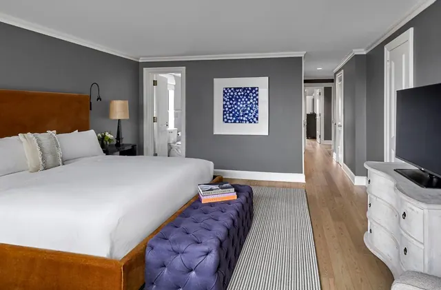 A bedroom with a large bed with an orange upholstered headboard and a purple tufted bench at its foot, a flat-screen television on a white dresser, and a bathroom visible through an open door.