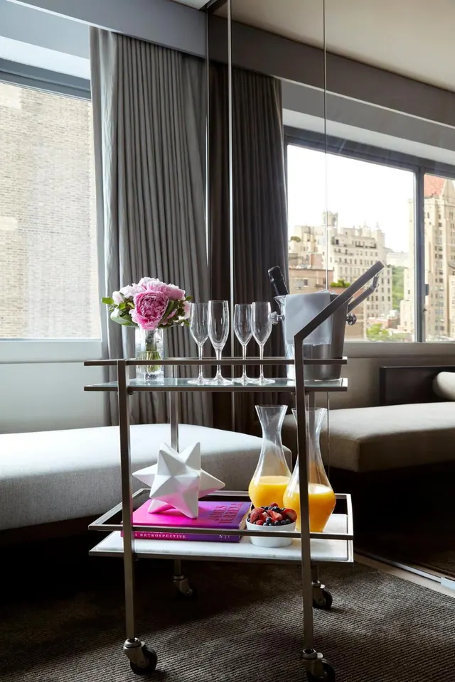 A bar cart holding flowers, champagne glasses, an ice bucket, juice carafes and a fruit bowl stands in a room with a view of city buildings through the window.