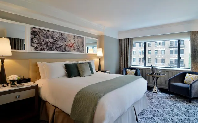 A bedroom with a king bed, framed artwork above the headboard, bedside lamps, and an armchair and round side table by a large window looking onto a brick building.