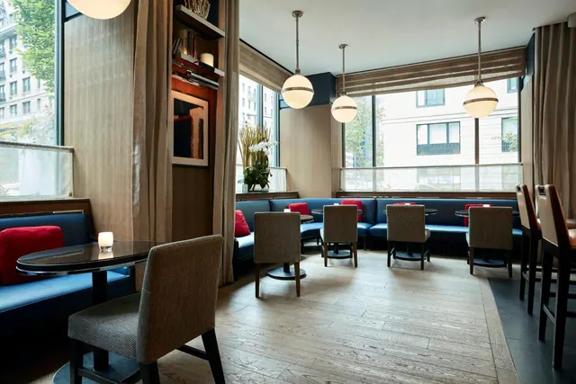 A lounge area with blue banquette seating, red cushions, wooden tables and chairs, and pendant lights, beside large windows looking onto a street with buildings.