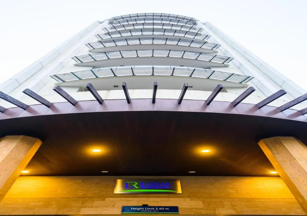 Low-angle view of a tall hotel tower with curved balconies and railings, a wood-clad entrance canopy with lights, and a height limit sign below.