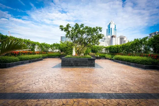 A paved garden terrace with a central planter holding a frangipani tree, trimmed hedges and red flowering shrubs, with tall city towers visible in the background under a partly cloudy sky.
