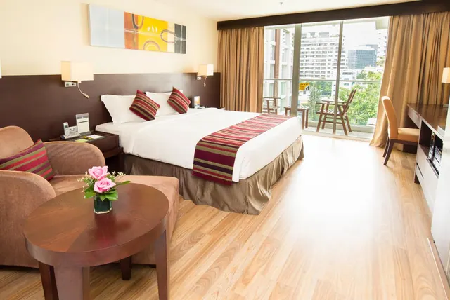 A hotel bedroom with a king bed, an armchair beside a round wooden table, a desk on the right, and sliding glass doors onto a balcony with city buildings beyond.