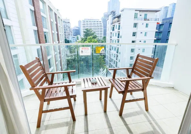 A balcony with two wooden chairs and a small wooden side table, with a glass railing and a view of high-rise buildings and trees.