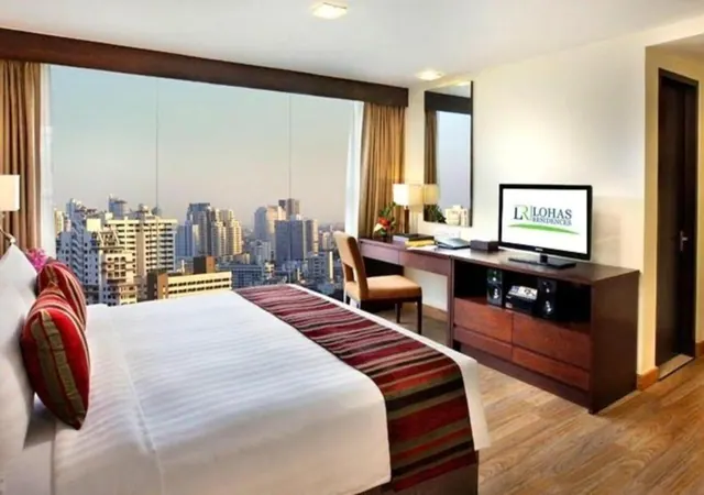 A bedroom with a king bed, a wooden desk with a flat-screen television, and a large window showing a city skyline of high-rise buildings.