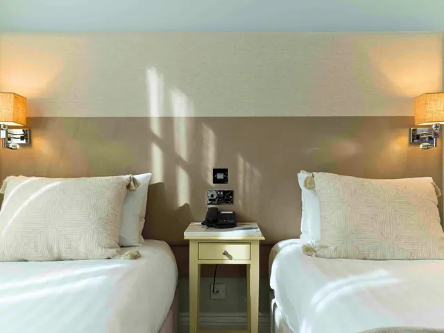 Two single beds with patterned cushions stand either side of a yellow bedside table holding a telephone, with wall lamps above each bed and sunlight falling across the headboard wall.
