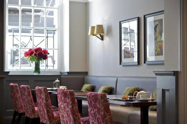 A dining room with pink paisley upholstered chairs at wooden tables, a grey banquette along the wall, a tall window with a vase of red flowers, and a teapot and pastry set on one table.