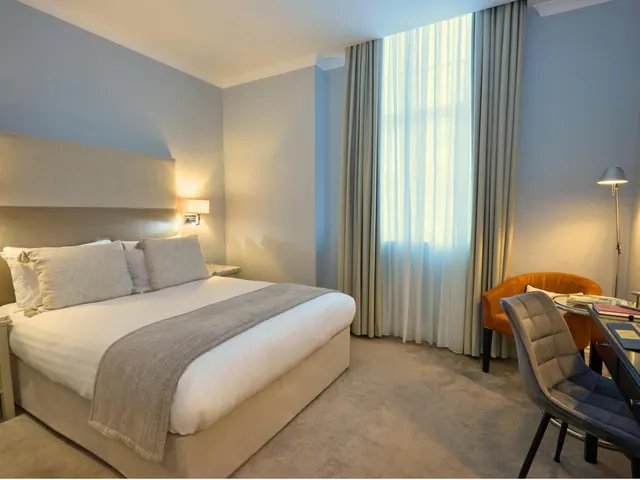 A bedroom with a double bed, an orange armchair, a grey chair at a round desk, and floor-length curtains beside a tall window.