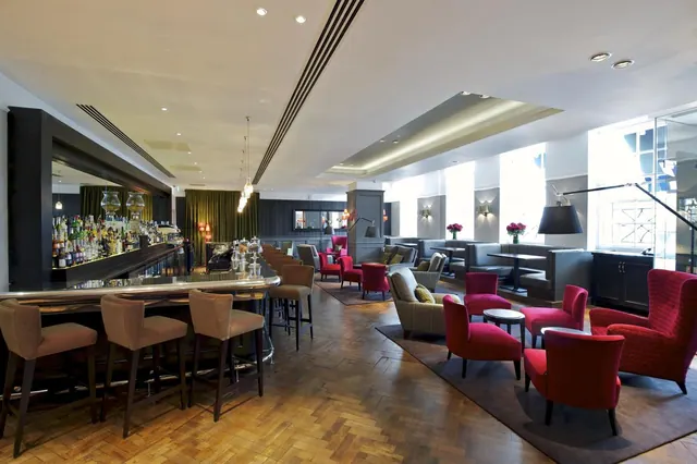 A lounge with a curved bar counter, bar stools, shelves of bottles, red armchairs and grey banquettes on a wooden parquet floor.