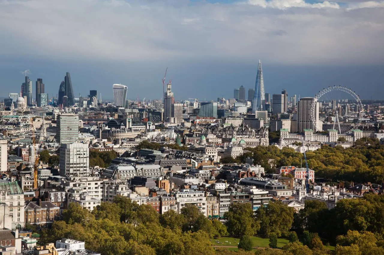 Elevated view over central London showing The Shard, the London Eye and office towers beyond rooftops and tree-lined parkland under a cloudy sky.